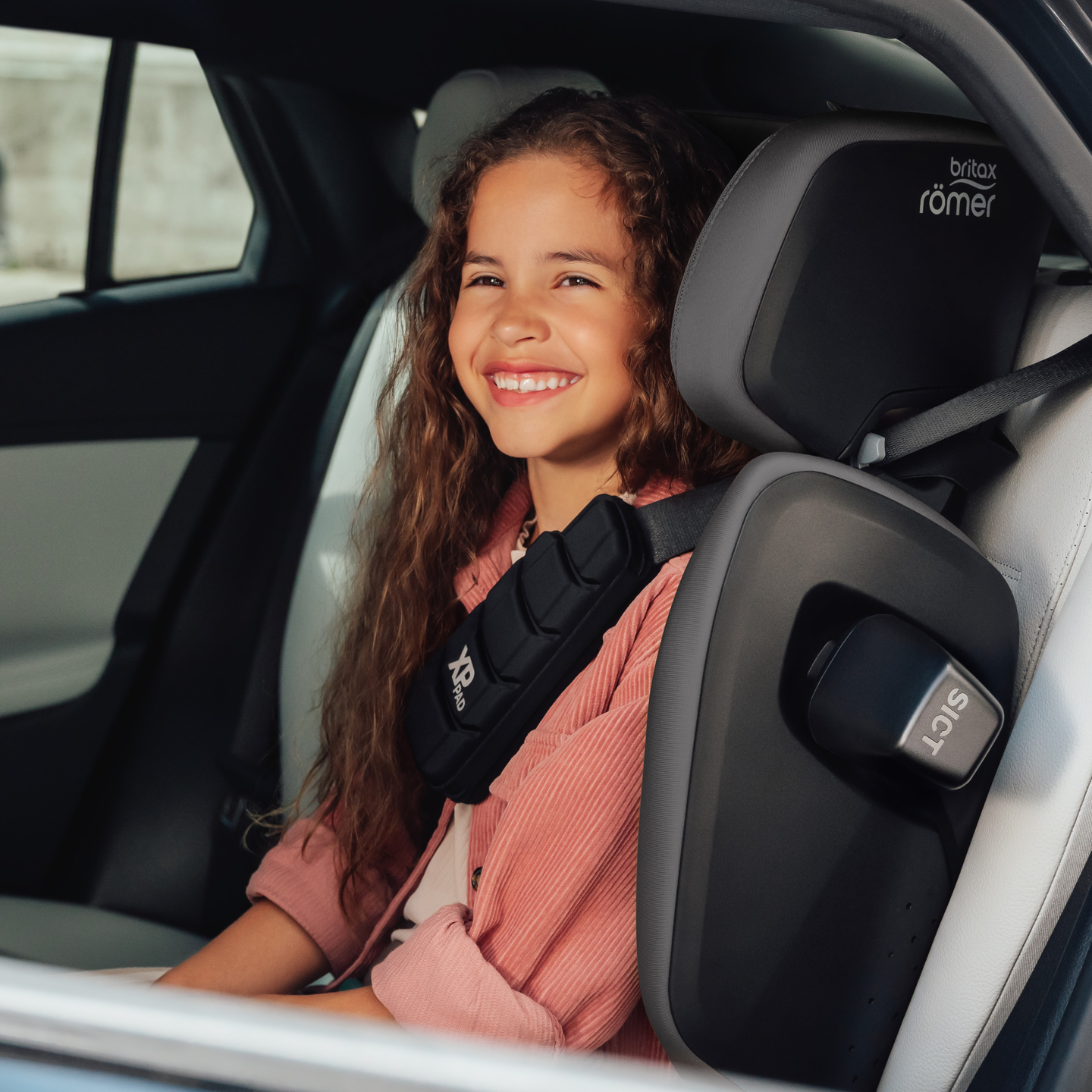 Leende barn i baks&auml;tet p&aring; en bil, s&auml;krat i en Britax R&ouml;mer KIDFIX PRO b&auml;ltesstol med h&ouml;g rygg.